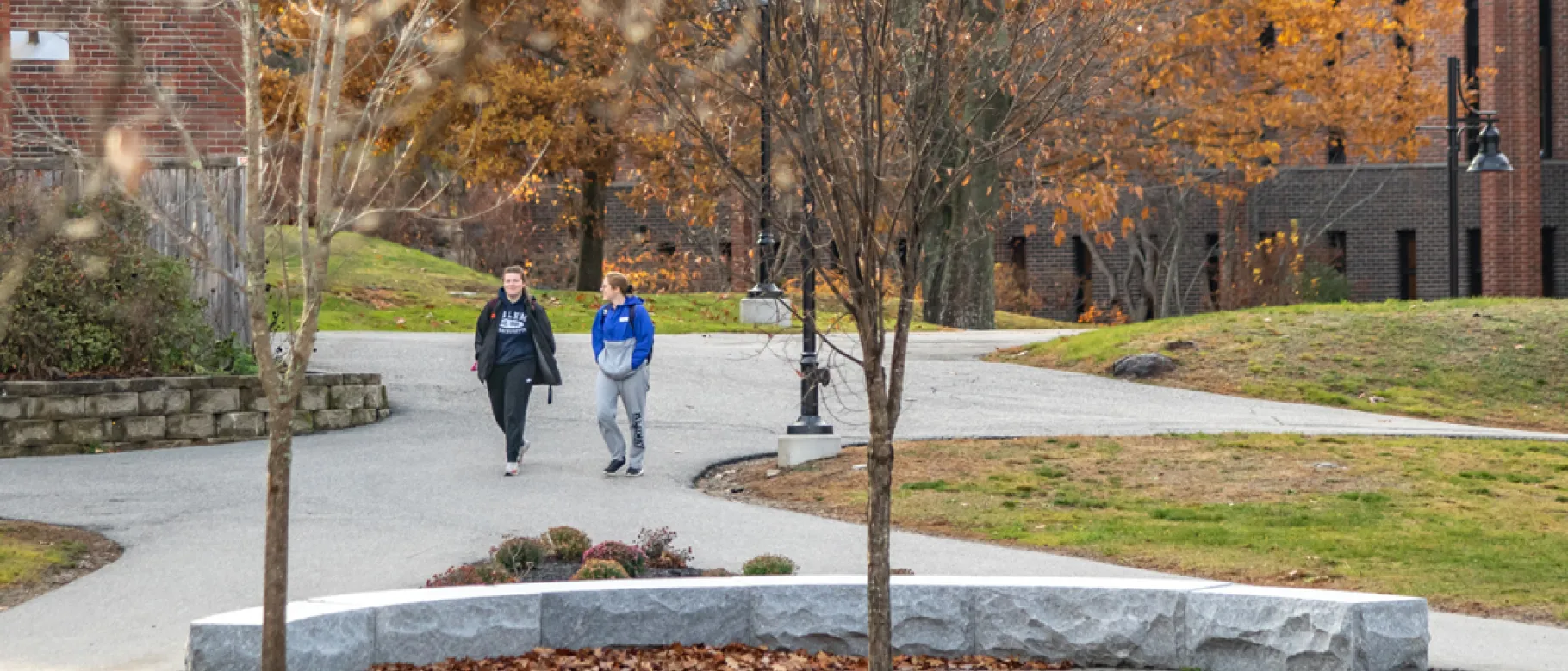 Two U N E students walk on a paved path through the greenery of Biddeford Campus
