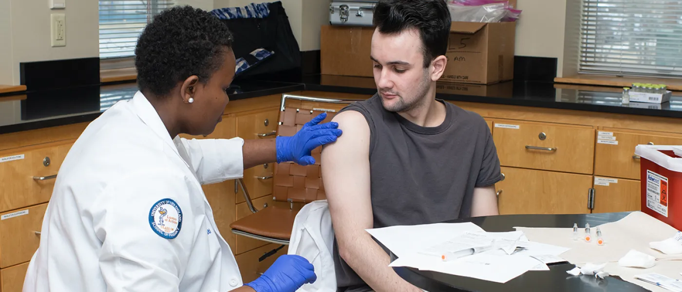 A pharmacy student practices giving a vaccine via needle to another student