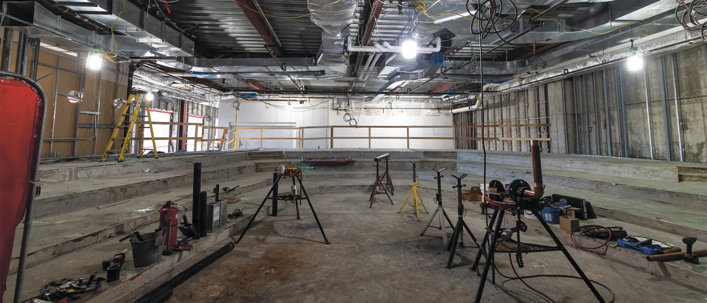 Construction site interior showing concrete floors, exposed ceiling infrastructure, work stands, and construction equipment during renovation.