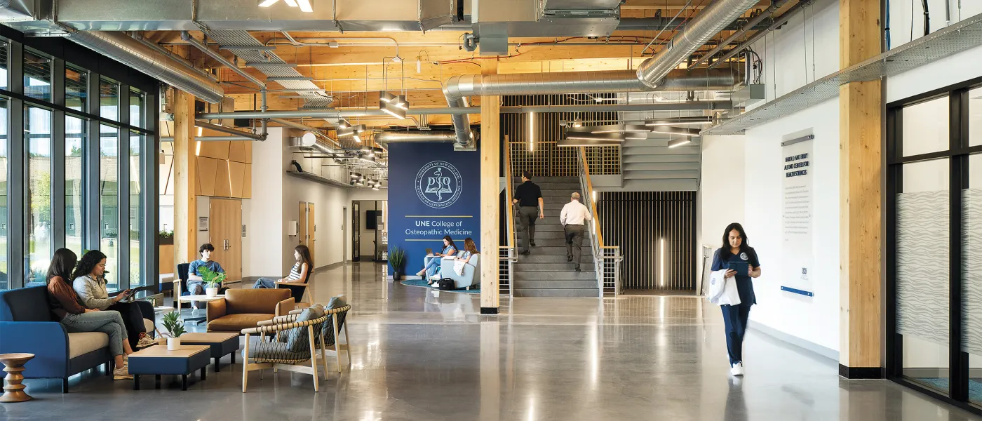 Modern UNE College of Osteopathic Medicine building interior with exposed wooden beams, polished concrete floors, central staircase, and students in a contemporary lounge area with natural light from floor-to-ceiling windows.