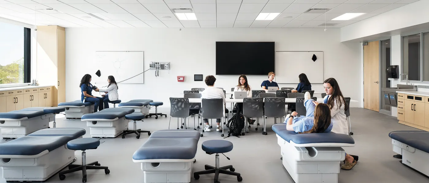 Clinical Skills lab with examination tables, medical equipment, students studying at tables, and wall-mounted displays.