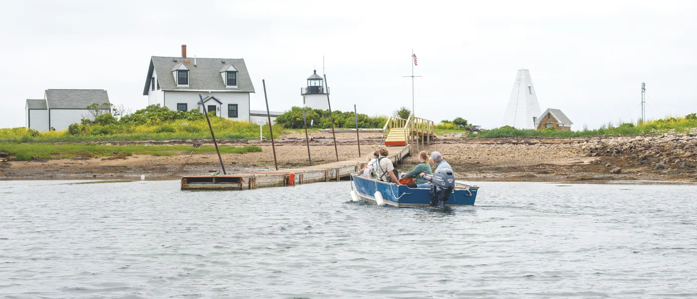 Students in a blue motorboat approach a wooden dock at a Maine coastal research station with a white keeper's house, lighthouse, and monument visible on the rocky shore.