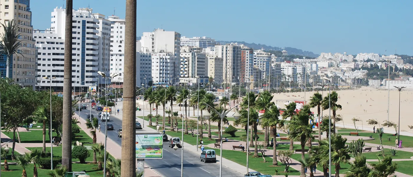 Coastal boulevard in Tangier with palm tree-lined streets, modern high-rise buildings, landscaped parks, and sandy beach along the Mediterranean Sea.