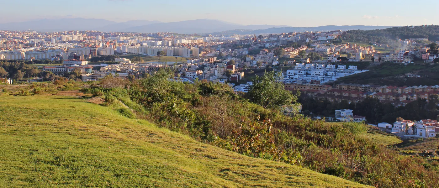 Panoramic hillside view of Tangier cityscape showing dense urban development stretching across rolling terrain with mountains visible in the distance.