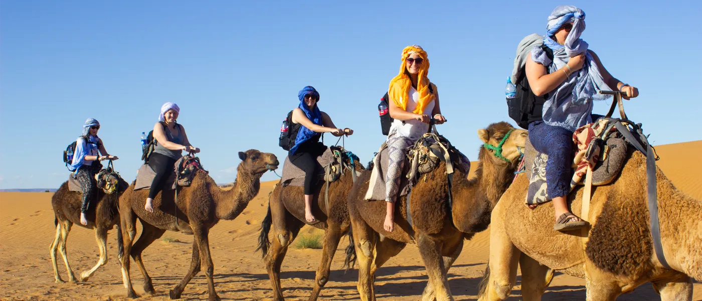 Five students wearing traditional head wraps ride camels in single file across Saharan sand dunes under clear blue sky.
