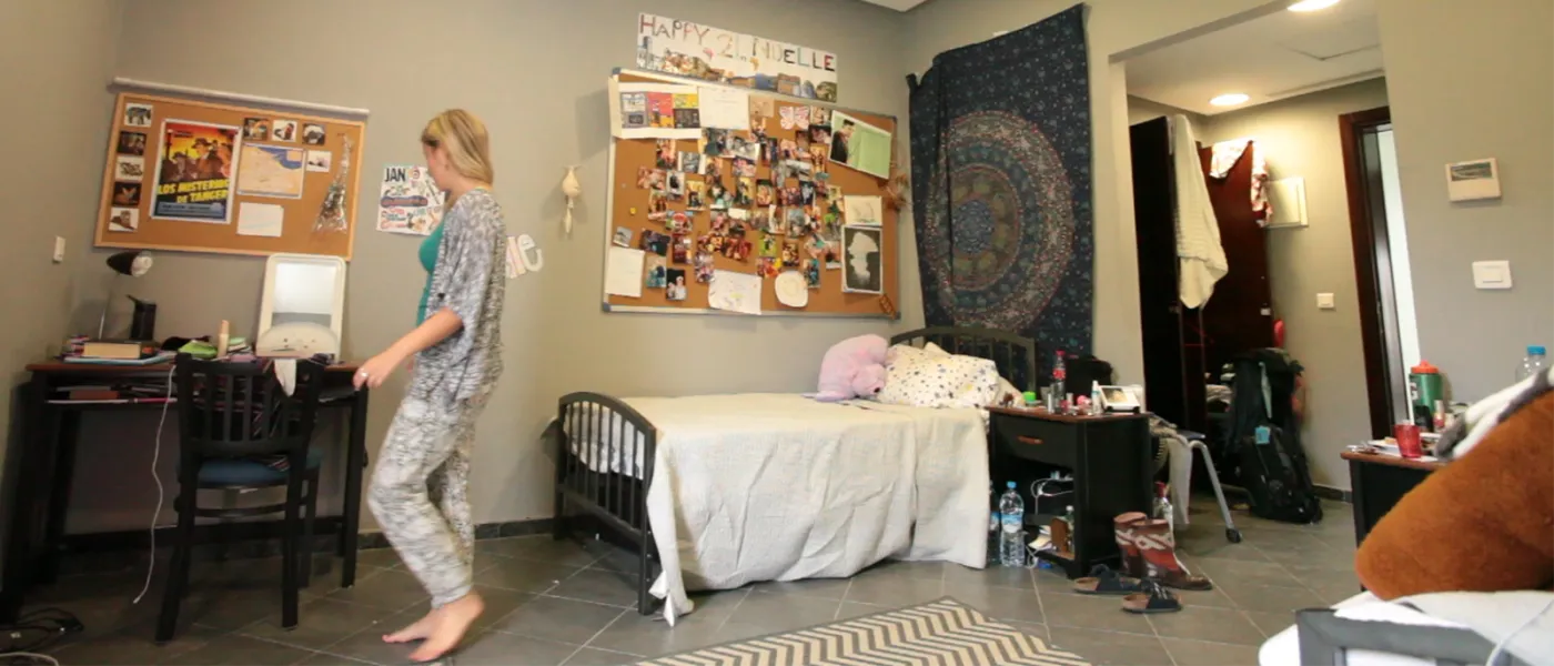 Student walks through dorm room with personal decorations, photo collages, tapestries, study desk, and patterned area rug.