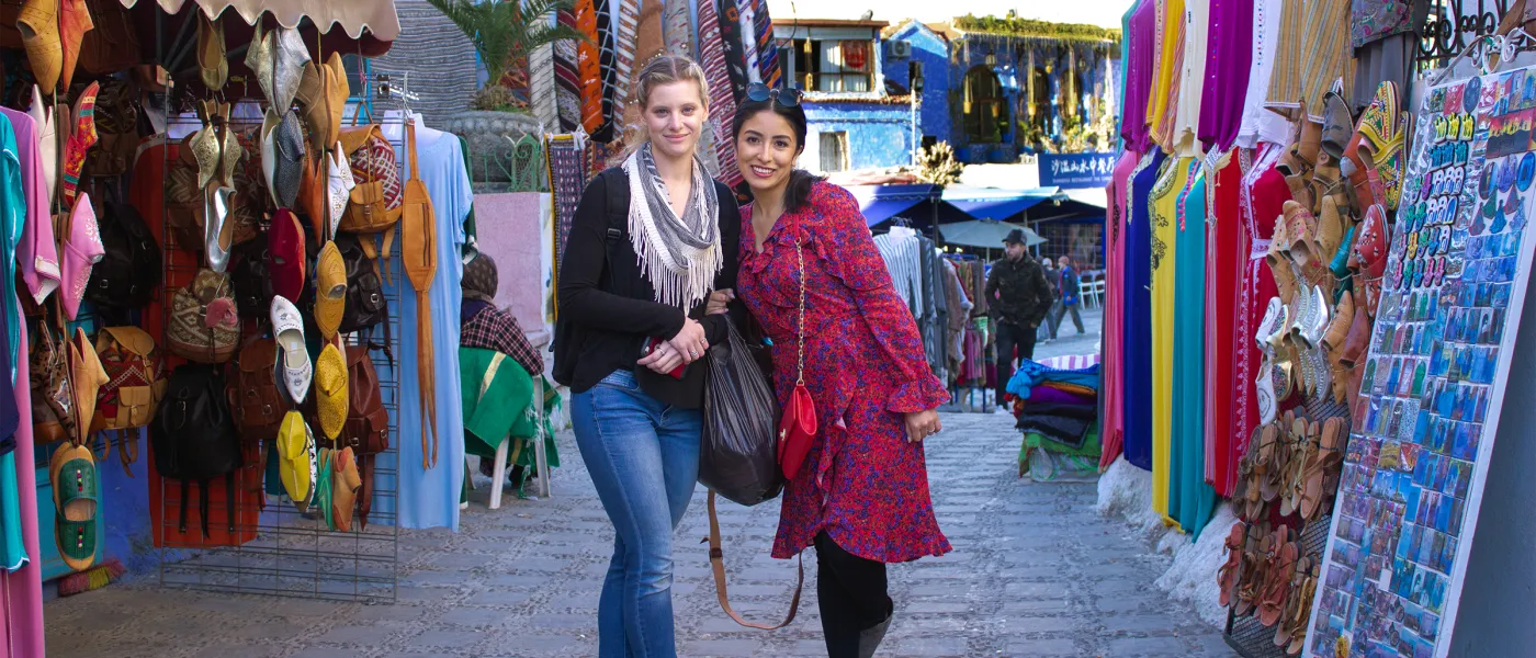 Two students walk arm-in-arm through vibrant marketplace with colorful leather bags, textiles, and crafts displayed on both sides.
