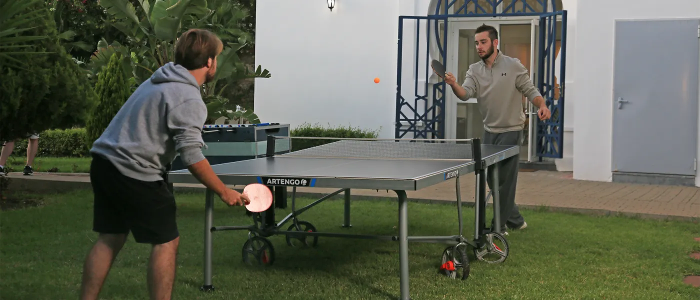 Two students play ping-pong on outdoor table in landscaped garden area at UNE Tangier Campus residence.