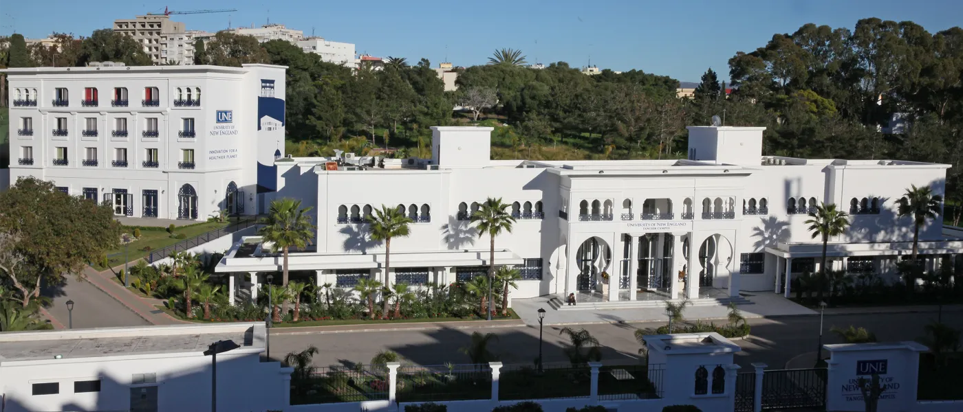 Aerial view of UNE Tangier Campus showing white buildings with Moroccan design, residence hall, landscaped grounds, and surrounding hillside neighborhood.