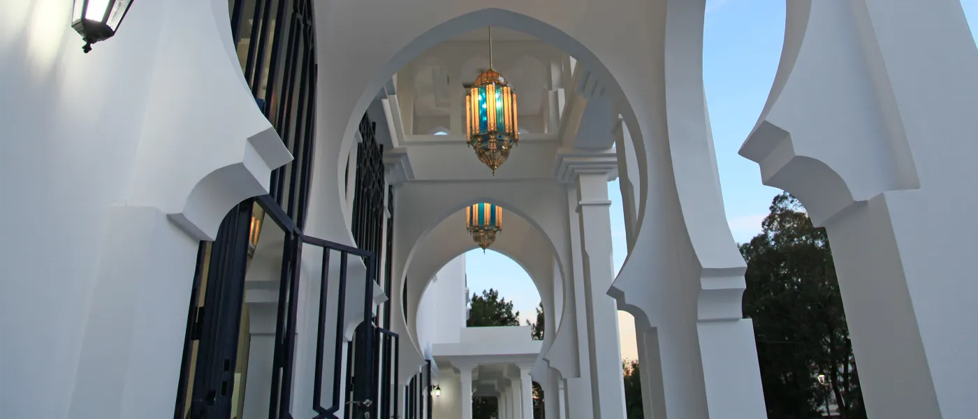 Covered outdoor corridor with white columns, horseshoe arches, decorative hanging lanterns, and traditional Moroccan architectural details.
