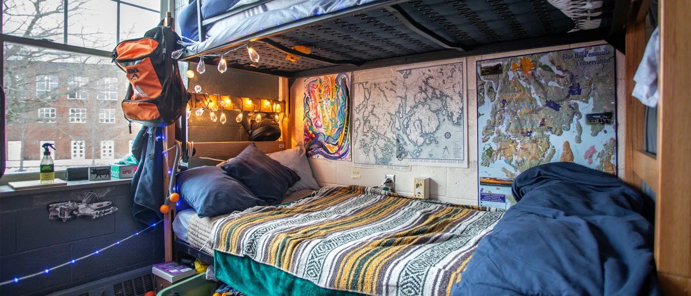 Lofted bed with string lights and a hanging backpack, decorated with topographic maps on the wall, a striped bedspread, and a window view of campus buildings.