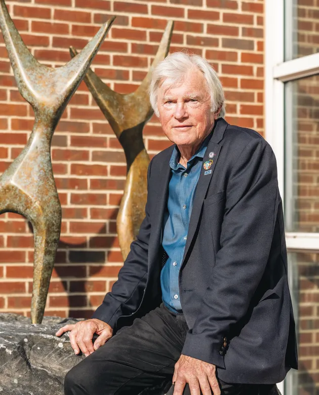 David Evans Shaw, HON '22, sits beside a bronze sculpture of three stylized dancing figures mounted on stone outside a brick UNE building, wearing a dark blazer over a blue shirt with a U N E pin