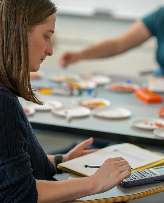 A student takes notes and uses a calculator during a math class.
