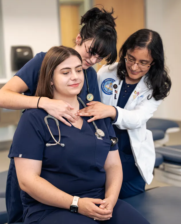 Two UNE College of Osteopathic Medicine students practice osteopathic manipulative medicine techniques on a classmate during a skills lab, with one student in a white coat applying hand placement to the seated student's shoulder.