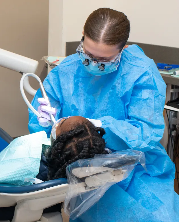 A dental student in a blue gown, mask, and loupes performs a procedure on a young patient in a dental chair during a community outreach event.