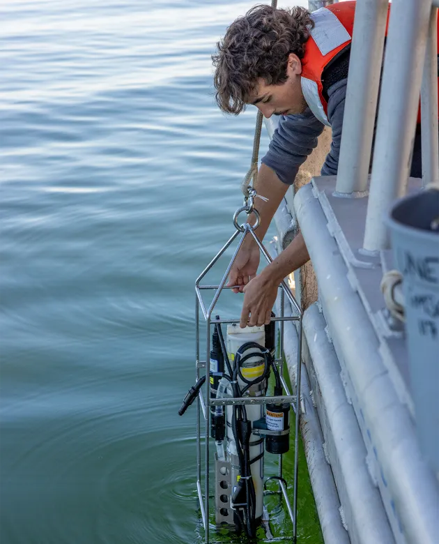 A UNE marine sciences student wearing an orange life jacket lowers a water sampling instrument over the side of a research vessel to collect data.