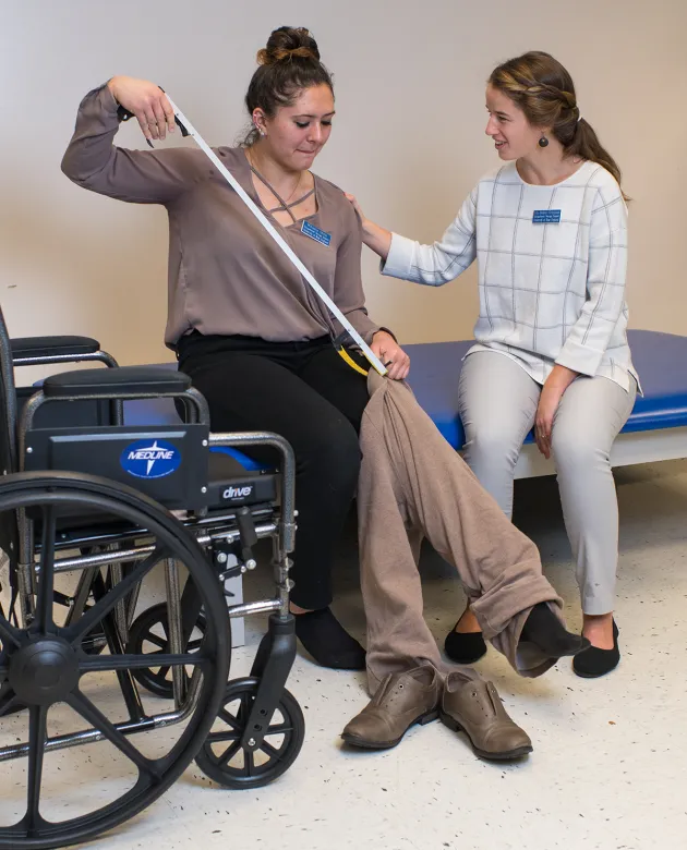 Two UNE occupational therapy students practice an adaptive dressing technique, with one student using a dressing stick to pull up pants while the other provides guidance and support during a skills lab.