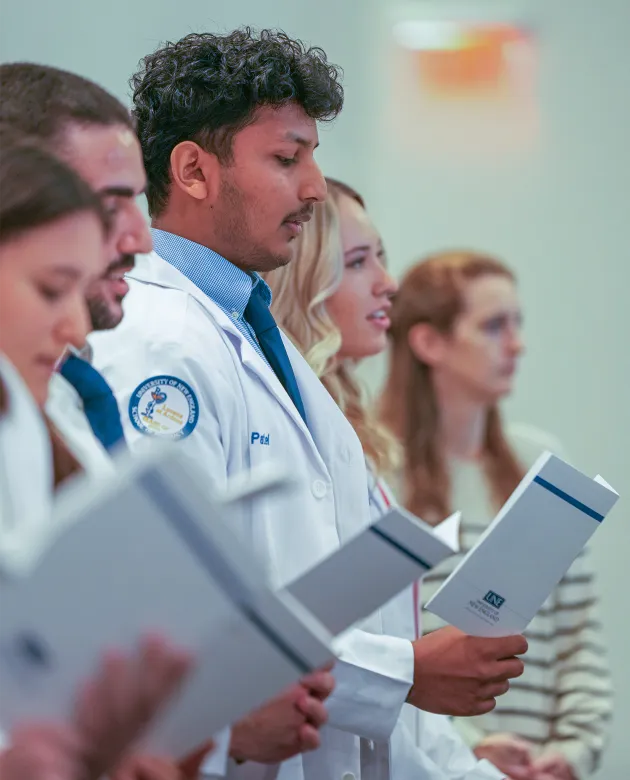 A row of UNE pharmacy students in white coats read aloud from programs during the oath recitation at a white coat ceremony.