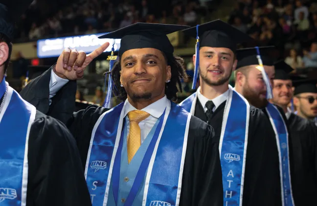 A student in academic regalia points to his black cap at graduation