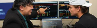 A business professor talks about finance to a student while pointing to dual monitors that display financial data