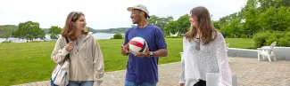 Three U N E students walk and chat together on a campus patio, with one carrying a volleyball. A lush green lawn, hammock, and the river are visible in the background.