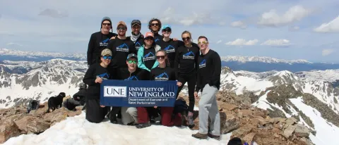 A group of students in winter gear stand behind a U N E banner on the top of Mt. Quandary