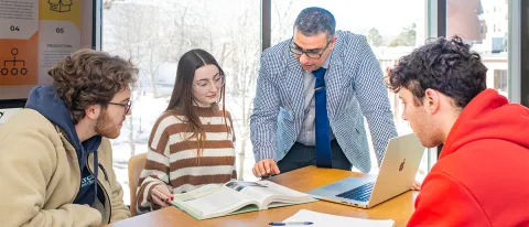 Three students review study materials with a professor at a large table