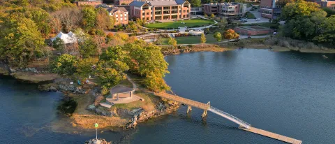 Aerial view of UNE's Biddeford campus in autumn, showing the waterfront campus with brick academic buildings, a small island with pavilion connected by a wooden footbridge, surrounded by colorful fall foliage and the Saco River estuary.