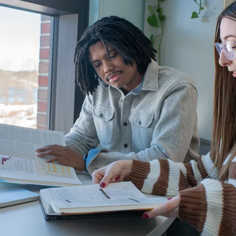 Two students read a textbook together in a room overlooking Biddeford Campus